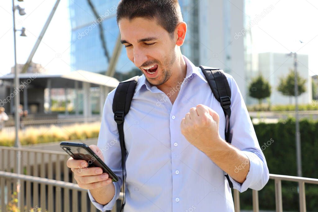 Excited young business man checking good news on smartphone. University student celebrating with mobile phone in his hand in modern city street.