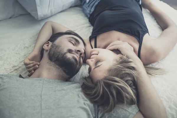 Loving lyckliga par i kärlek leende och kyssa hennes kind på sängen, Stockfoto