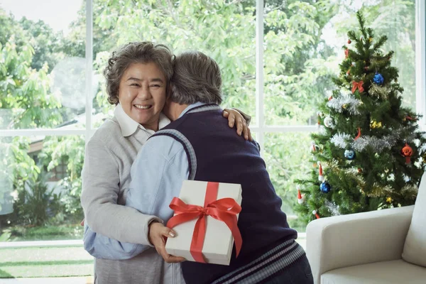 幸せなカップル高齢者の男性と女性の抱き合いとギフトボックスを保持し 彼らは家で一緒に笑顔で満足しています 退職と医療ライフスタイルと恋人関係の概念 ストックフォト