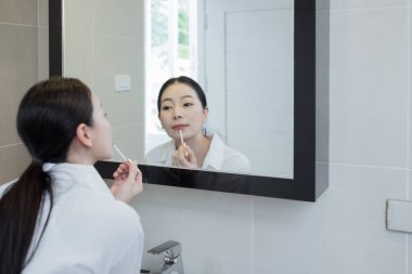 Asyalı kadın beyaz gömlek giyip makyaj yapıyor. Aynanın önünde, banyoda.