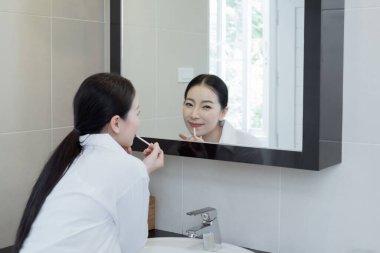 Beyaz gömlekli, makyajlı, aynanın karşısında gülümseyen Asyalı bir kadın.