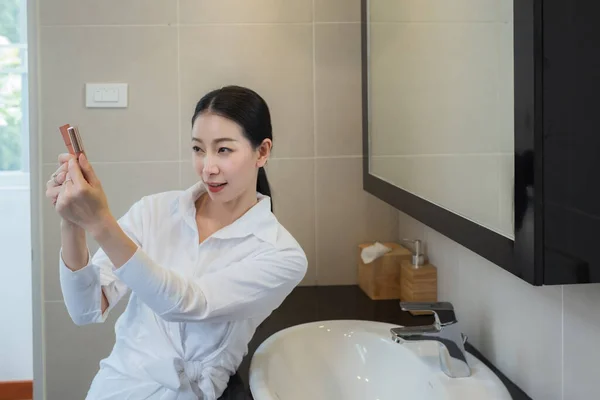 亚洲女人穿着白衬衫 在浴室的镜子前选择化妆 — 图库照片