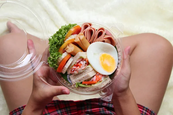 Salata, sahanda yumurta, pastırma, domates yuvarlak plastik bir kutuda. Bu, bir kadının elinde sergilenen çevrimiçi bir mağazadan sipariş edilmiş. Bir kadının elinde sergilenen çevrimiçi bir mağazadan alınmış bir sipariş..