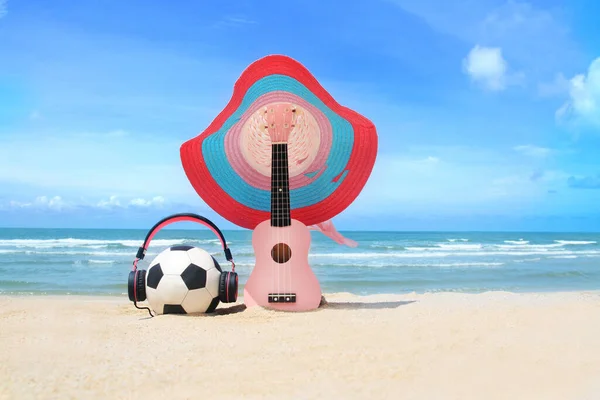 Pembe ukulele, şapka, kulaklık, futbol mavi gökyüzünde ve plaj arka planında 