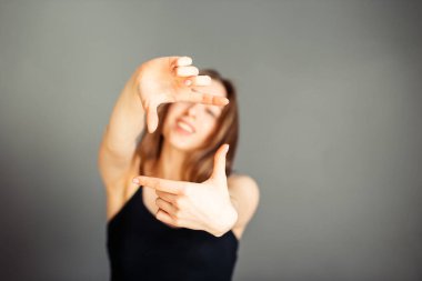 Saçları siyah bluzlu bir kız elleriyle fotoğraf çekiyor. Gri bir arka planda. Makyajsız, rötuşsuz.