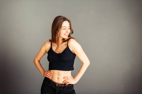 Девушка в черном топе с раскинутыми на бедрах волосами, смотрит в сторону и улыбается на сером фоне. Без макияжа, без ретуши
.