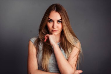 Gri tişörtlü, çenesini eliyle tutarak gri duvardaki çerçeveye bakan güzel sarışın bir kadının stüdyo portresi.