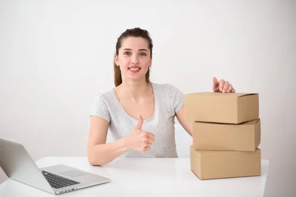 A girl with a pleasant smile on a light background sits at a table with a computer and corton boxes and shows thumb up. delivery service concept
