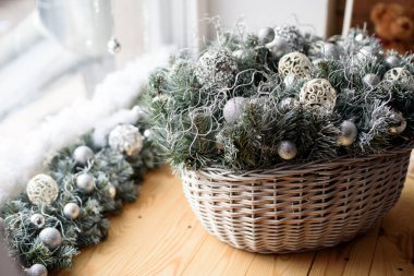 İçinde Noel ağacı dalları ve gümüş oyuncaklar olan sepet ön taraftaki ahşap pencere pervazında, bir pencerenin arka planında.