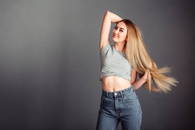 Gri bluzlu, kot pantolonlu, sarışın, eli başının arkasında olan ve saçlarını siyah bir duvara dayayan zayıf bir kız.