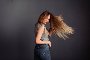 Gri bluzlu, kot pantolonlu, sarışın, eli başının arkasında olan ve saçlarını siyah bir duvara dayayan zayıf bir kız.