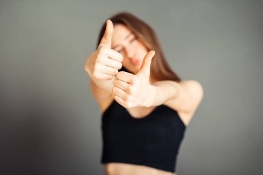 Saçları siyah bluzlu kız başparmağını kaldırıyor. Gri arka planda mutlu. Makyajsız, rötuşsuz.