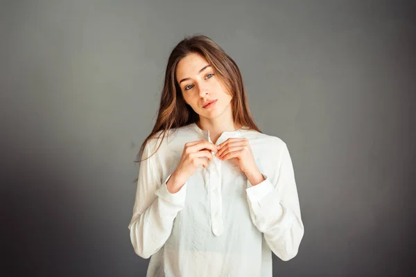 Serious girl with loose hair on her head in a light blouse. hands near the collar. Portrait. On a gray background. Without makeup, without retouching.