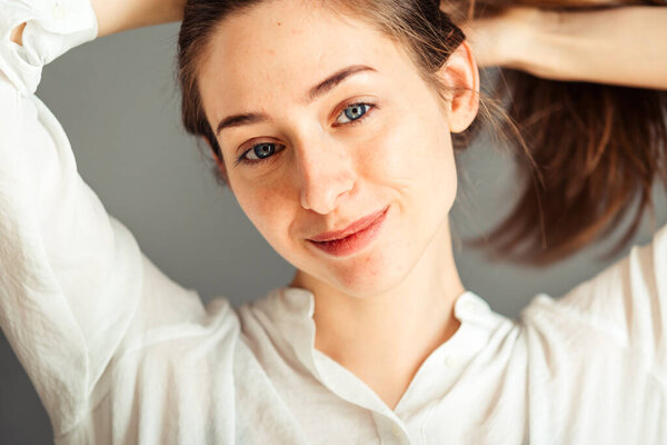 Улыбающаяся девушка с довольным лицом держит волосы руками на сером фоне крупным планом. Обнажённый макияж, без ретуши
.