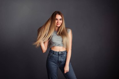 Gri bluzlu, kot pantolonlu, sarışın, eli başının arkasında olan ve saçlarını siyah bir duvara dayayan zayıf bir kız.