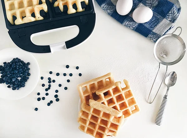 Making Belgian waffles at home - waffle iron, kitchenware and ingredients - fresh blueberry, eggs and flour. Cooking background. Flat lay, top view, copy space