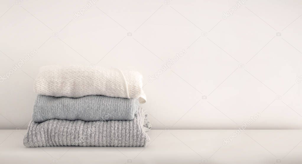 Stack of neatly folded white, grey and blue tone woolen knitwear. Minimal lifestyle, capsule wardrobe. Autumn-winter wardrobe concept. Light and airy, horizontal, copy space