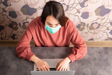 Yüz maskesi takan ve evde dizüstü bilgisayar kullanan öğrenci görüntüsü.