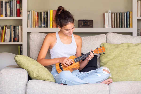 Ukulele oynayan genç bir kız evdeki kanepede oturuyor.