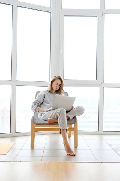 Woman in armchair working on laptop near panoramic window