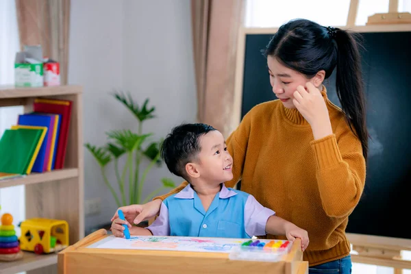 在幼儿园的课堂上 亚洲老师教她的学生用彩色铅笔在有背边背景的桌子上画画 这个图像可以用来作为返回学校和接受教育的概念 — 图库照片