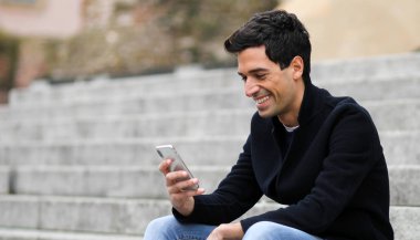 Açık merdivenlerde oturan bir akıllı telefon kullanan genç adam