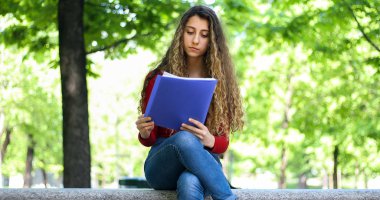 Güzel bir üniversite öğrencisi parkta bir bankta kitap okuyor.