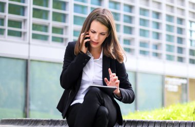 Dışarıda dijital tablet kullanan gülümseyen iş kadını bankta oturuyor ve telefonda konuşuyor.