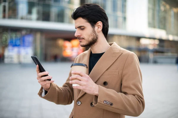 Mladý Muž Pomocí Svého Mobilu Drží Šálek Kávy Při Procházce — Stock fotografie