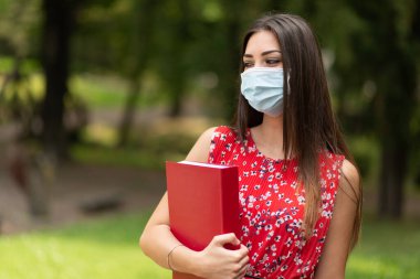 Maskeli kadın öğrenci parkta kitap tutuyor.