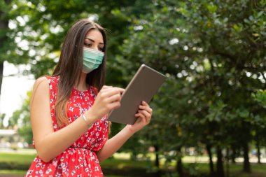 Maskeli genç bir kadın parkta tablet kullanıyor.