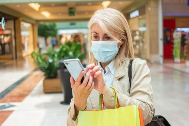 Alışveriş merkezinde yürürken cep telefonunu kullanan maskeli beyaz bir kadın.