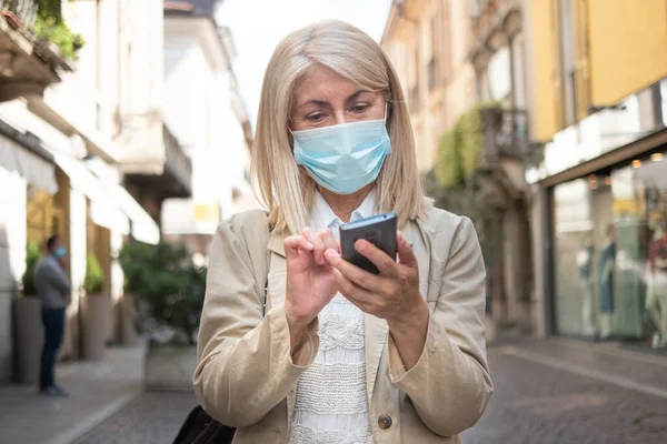 コロナウイルスのパンデミック時にスマートフォンを使いながら街を歩く熟女 — ストック写真