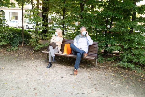 Man talking on the phone while keeping his distance from a woman on the park bench, coronavirus safety and social distancing concept