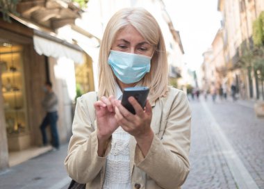 Koronavirüs salgını sırasında şehirde yürürken cep telefonu kullanan maskeli olgun sarışın kadın.