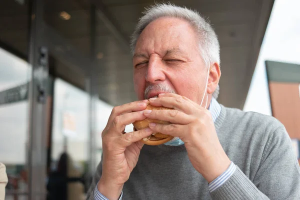 男食べる彼のファーストフードランチハンバーガー中にマスクで彼のあご コロナウイルスの概念 — ストック写真