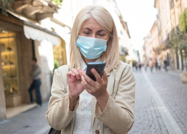 マスク成熟したブロンドの女性を使用して彼女の携帯電話のスマートフォン中に街を歩く中コロナウイルスパンデミック — ストック写真