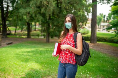 Maskeli kadın öğrenci parkta kitap tutuyor.