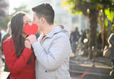 Genç çift öpüşüyor ve kendilerini kalp şeklinde bir kartonla örtüyorlar.