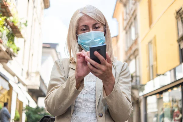 Masked Mature Blonde Woman Using Her Mobile Smartphone While Walking — Stock Photo, Image