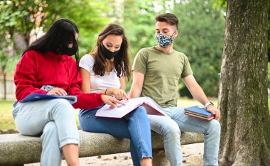 Üç öğrenci, Coronavirus zamanlarında bankta oturup maske takarak ders çalışıyor.