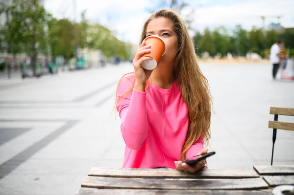 スマホを使いながら屋外でコーヒーを飲む幸せな若い女性 — ストック写真