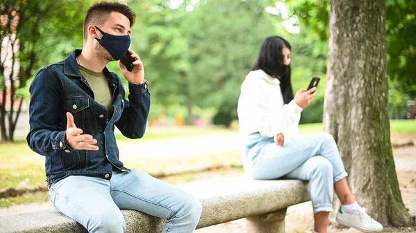 Young people keeping social distancing due to coronavirus while using their phone outdoor in a park