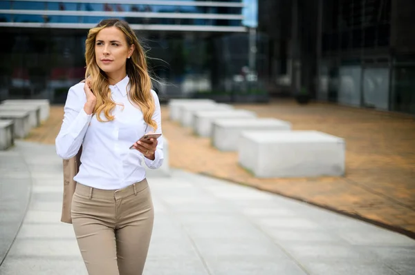 若い女性マネージャーはスマートフォンを使いながら屋外を歩く — ストック写真