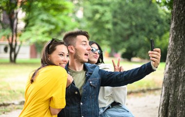 Parkta gülümseyen neşeli arkadaşlar bankta oturup akıllı bir telefonla selfie çekiyorlar.