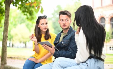 Üç öğrenci dışarıda bir bankta dijital tabletle birlikte ders çalışıyor.