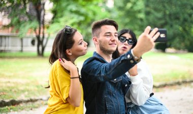 Parkta gülümseyen neşeli arkadaşlar bankta oturup akıllı bir telefonla selfie çekiyorlar.