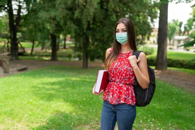 Parkta yürürken elinde kitap tutan maskeli kadın öğrenci, Coronavirus covid-19 eğitim konsepti.