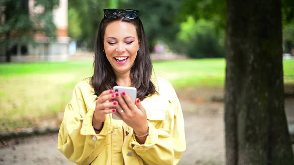 迷人的年轻女子在公园里用她的智能手机笑着 — 图库照片