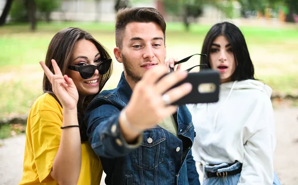 ベンチに座って スマートフォンを使って自撮りしている公園の陽気な笑顔の友人 — ストック写真